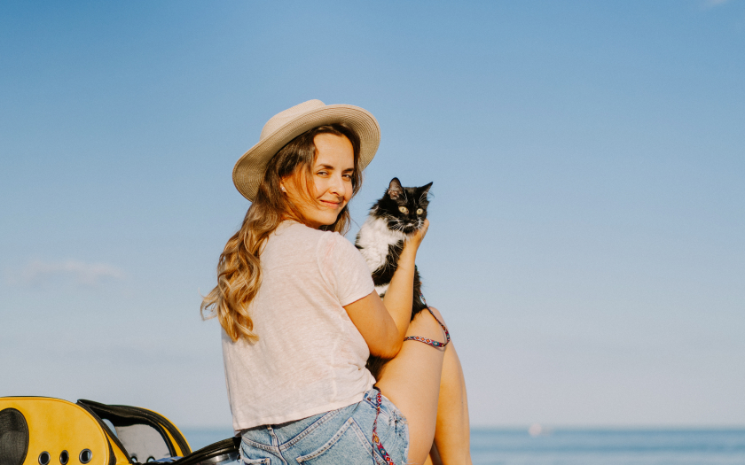 una mujer con sombrero y polera blanca sentada en una playa, sosteniendo a su gato que acaba de sacar de su mochila transportadora para viajar con mascotas.